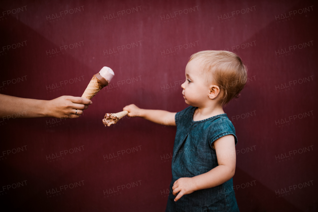 A side view of cute small toddler girl outdoors in summer, eating ice cream.