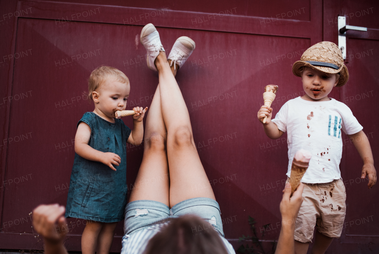 A young unrecognizable mother with two toddler children outdoors in summer, eating ice cream.