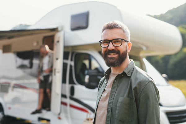 A portrait of happy man standing by car outdoors in campsite, looking at camera. Caravan family holiday trip.