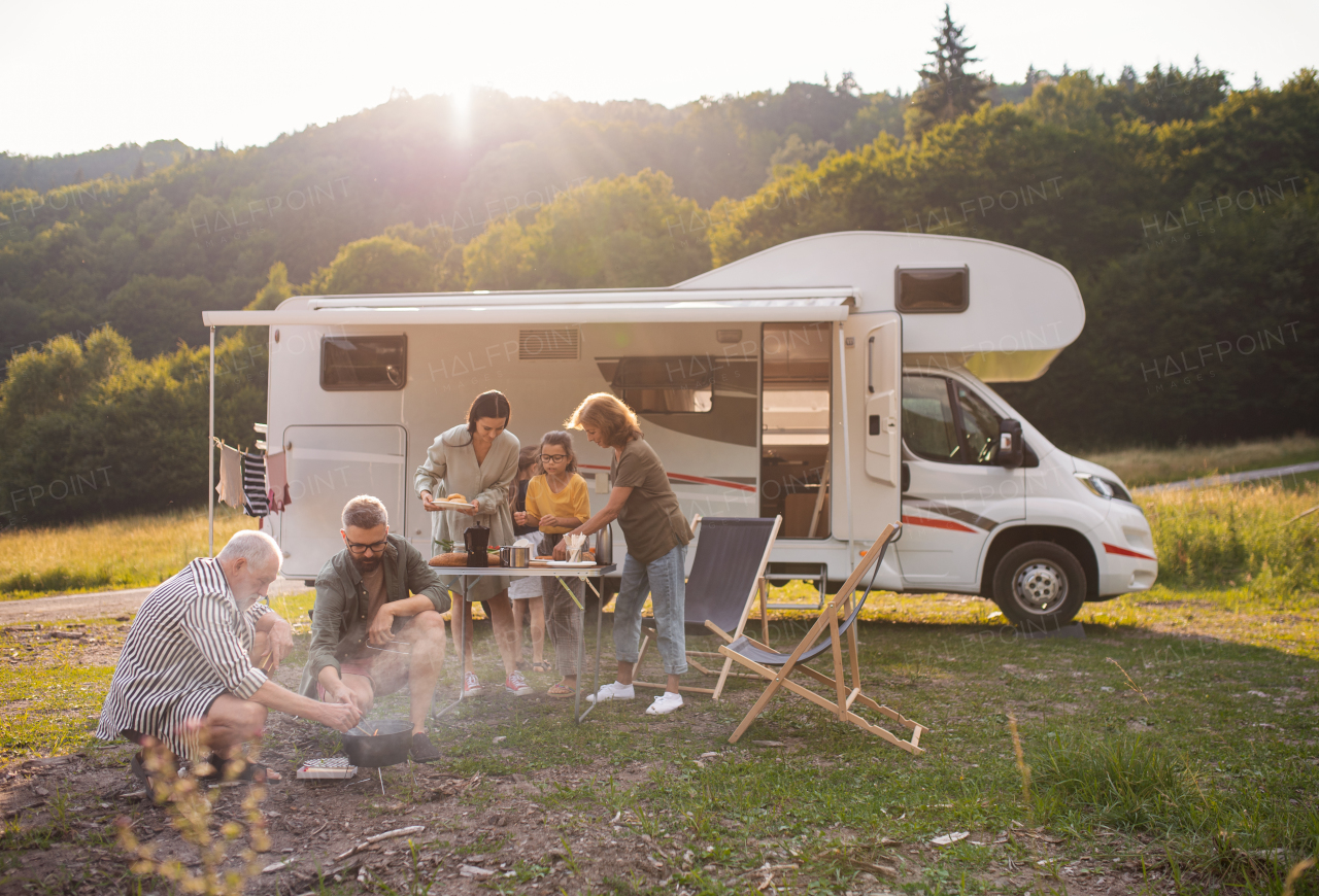 A multi-generation family sitting and eating outdoors by car, caravan holiday trip.