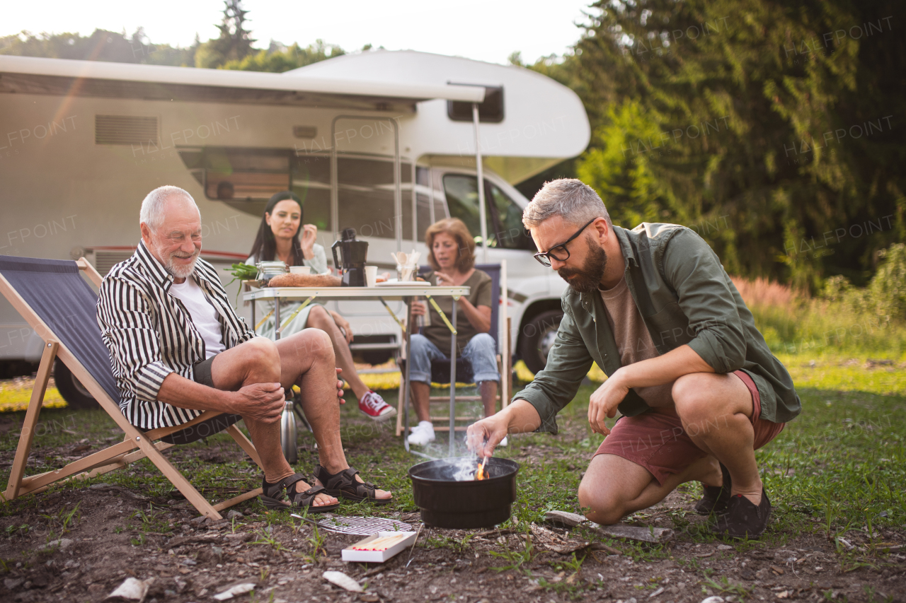 A multi-generation family preparing food on grill by car, caravan holiday trip.