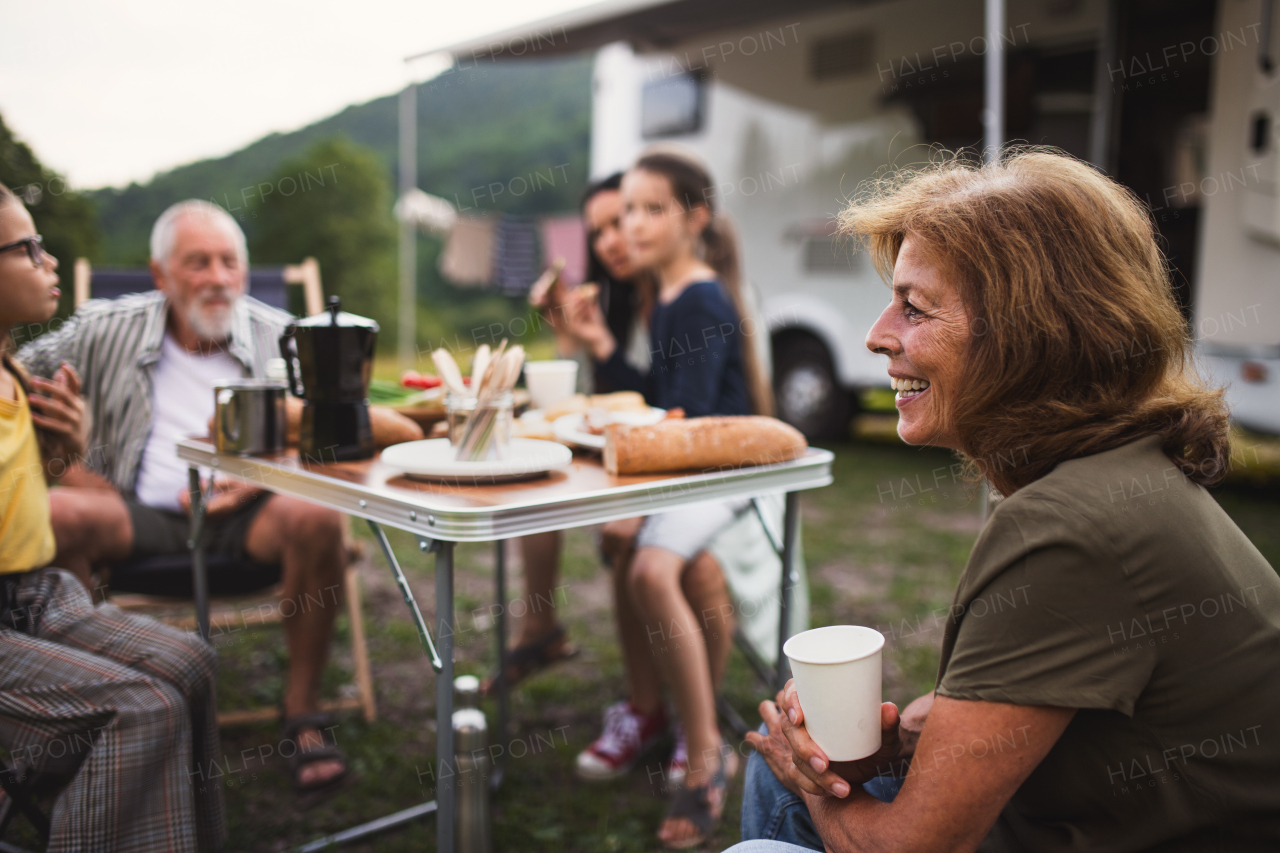 A multi-generation family sitting and eating outdoors by car, caravan holiday trip.