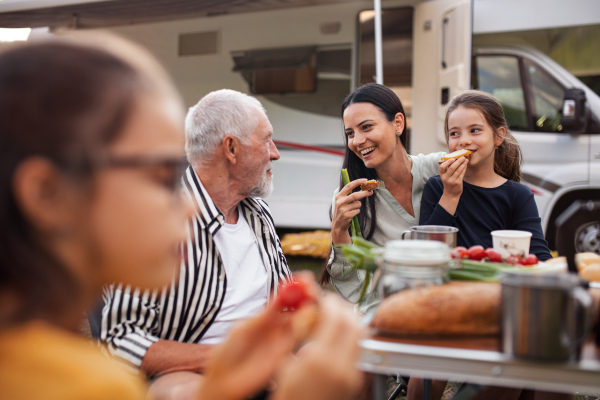 A multi-generation family sitting and eating outdoors by car, caravan holiday trip.