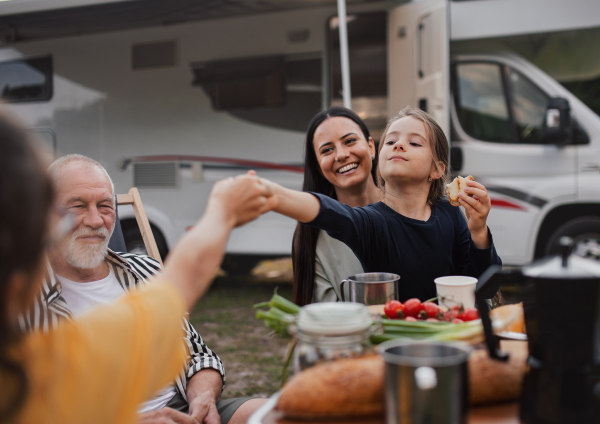 A multi-generation family sitting and eating outdoors by car, caravan holiday trip.