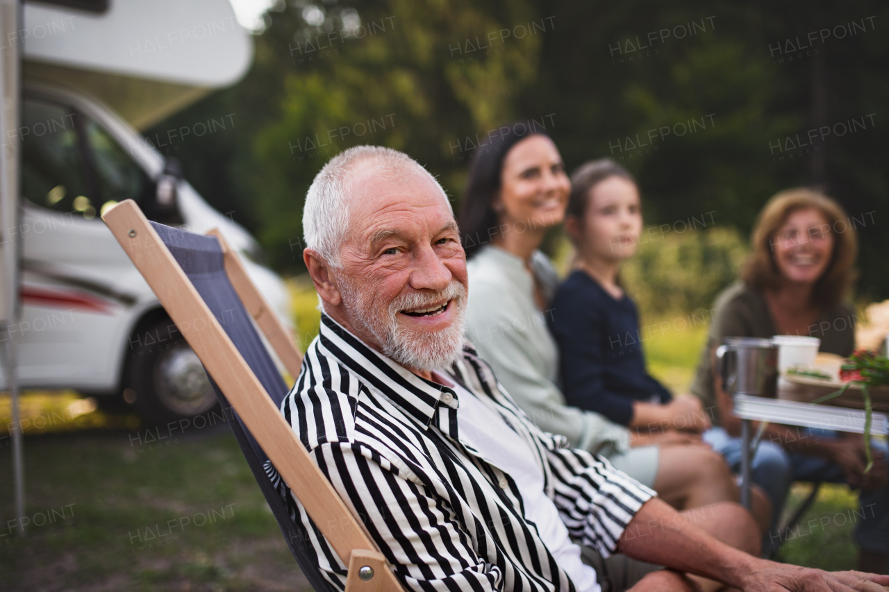 A senior man with multi-generation family sitting by car and looking at camera, caravan holiday trip.