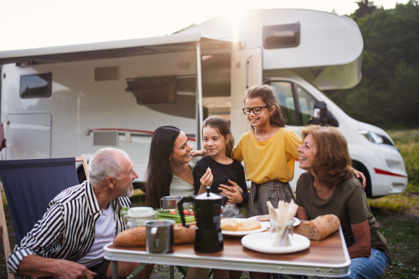 A multi-generation family sitting and eating outdoors by car, caravan holiday trip.