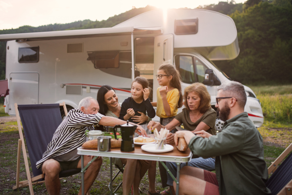 A multi-generation family sitting and eating outdoors by car, caravan holiday trip.