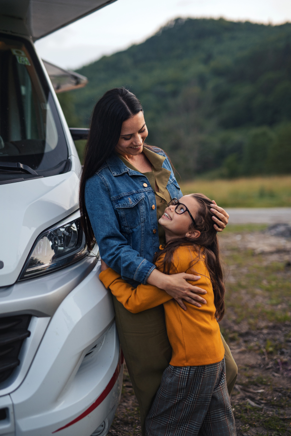 A mother with small daughter hugging by car outdoors in campsite at dusk, caravan family holiday trip.