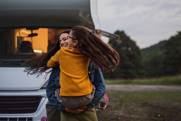 A happy mother with small daughter hugging by car outdoors in campsite at dusk, caravan family holiday trip.