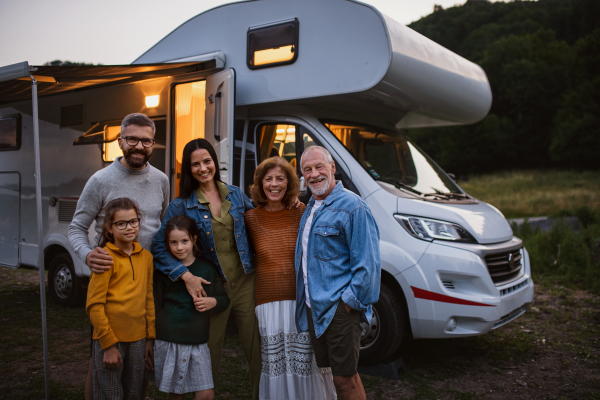 A portrait multi-generation family looking at camera and smilingoutdoors at dusk, caravan holiday trip.