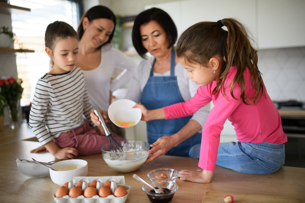 Happy small girls with mother and a grandmother making pancake mixture indoors at home, cooking.