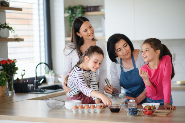 Happy small girls with mother and a grandmother making pancake mixture indoors at home, cooking.