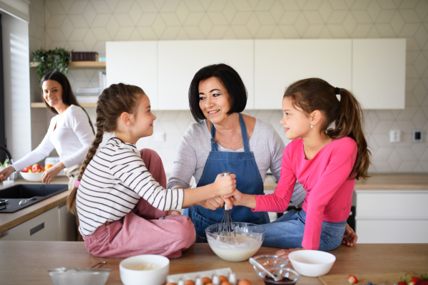 Happy small girls with mother and a grandmother making pancake mixture indoors at home, cooking.