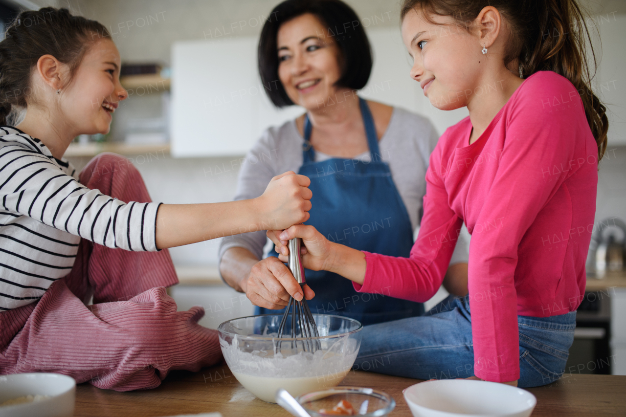 Happy small girls with a grandmother making pancake mixture indoors at home, cooking.