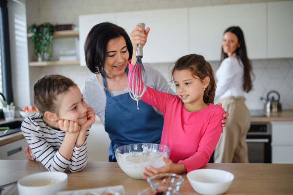 Happy small girls with mother and a grandmother making pancake mixture indoors at home, cooking.