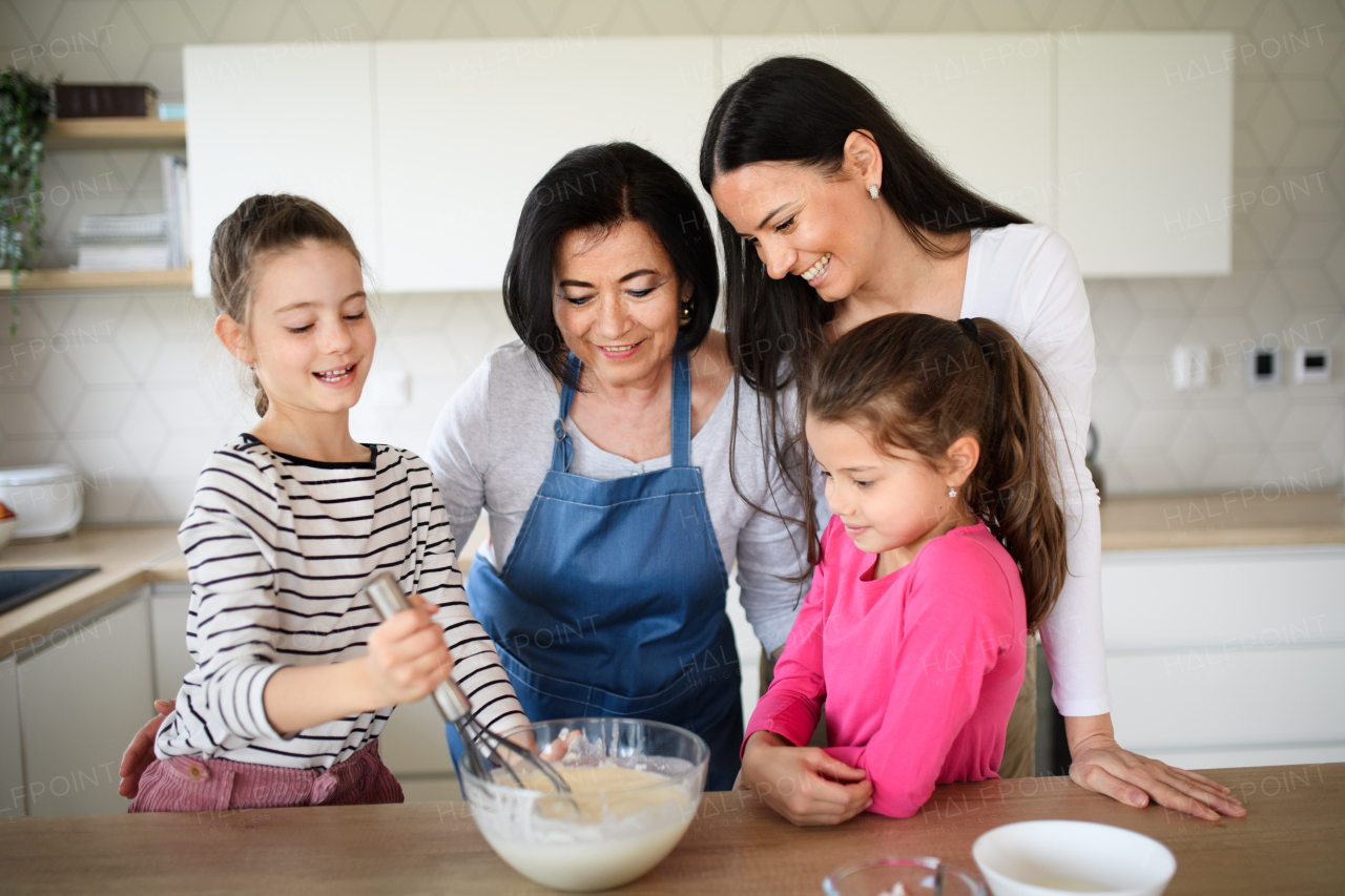 Happy small girls with mother and a grandmother making pancake mixture indoors at home, cooking.