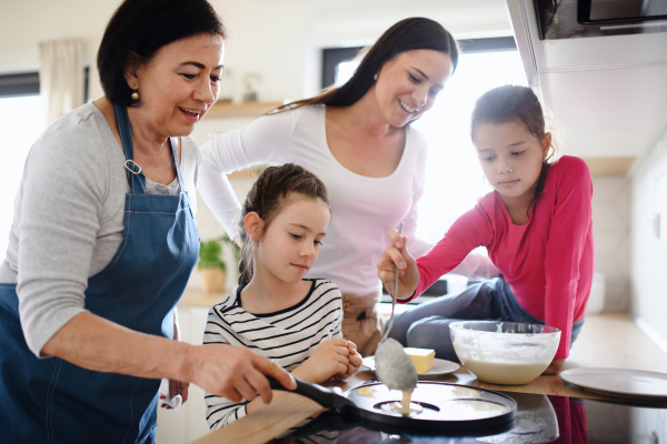 Happy small girls with mother and a grandmother making pancakes indoors at home, cooking.