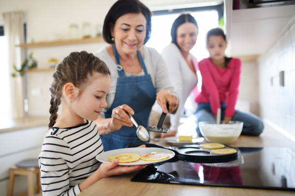 Happy small girls with mother and a grandmother making pancakes indoors at home, cooking.