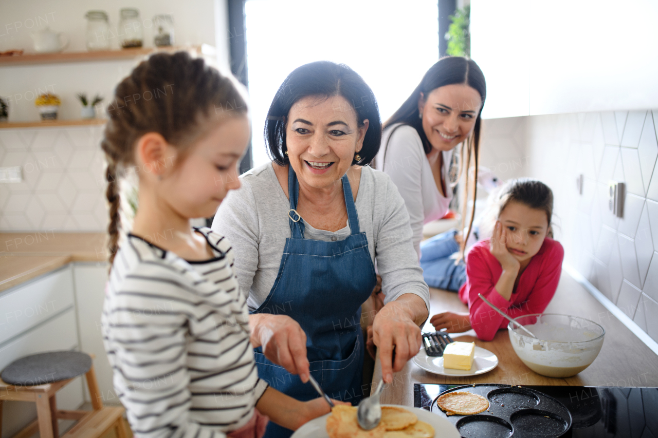 Happy small girls with mother and a grandmother making pancakes indoors at home, cooking.