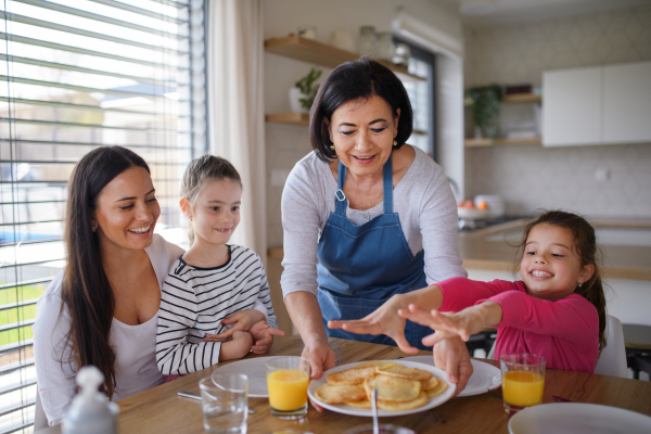 Happy small girls with mother and grandmother eating pancakes indoors at home, having a lunch.