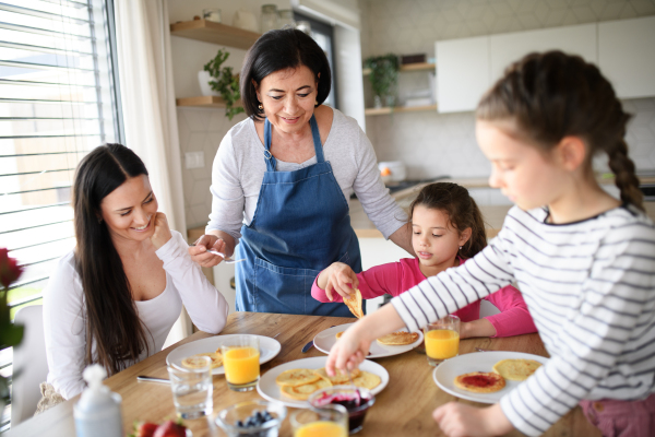 Happy small girls with mother and grandmother eating pancakes indoors at home, having a lunch.