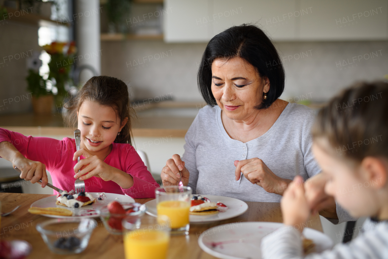 Happy small girls with a grandmother eating pancakes indoors at home, having lunch.