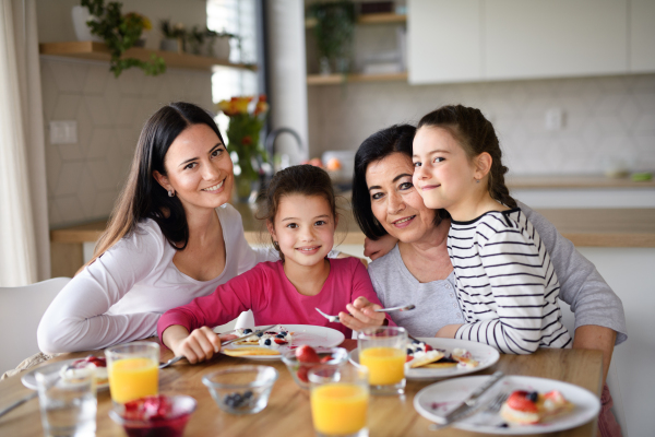 Happy small girls with a mother and grandmother eating pancakes indoors at home, looking at camera.