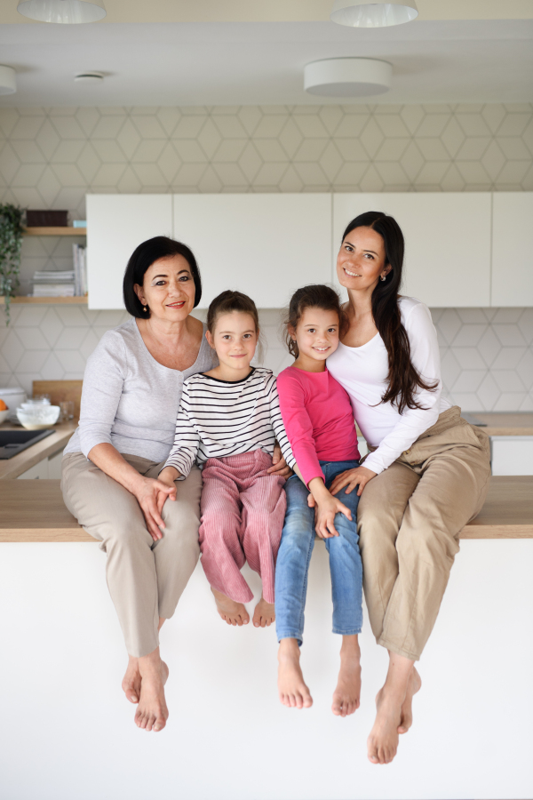 Portrait of happy small girls with mother and grandmother sitting on kitchen counter indoors at home, looking at camera.