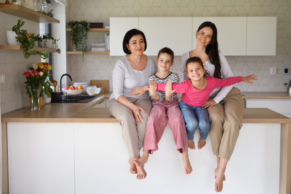 Portrait of happy small girls with mother and grandmother sitting on kitchen counter indoors at home, looking at camera.