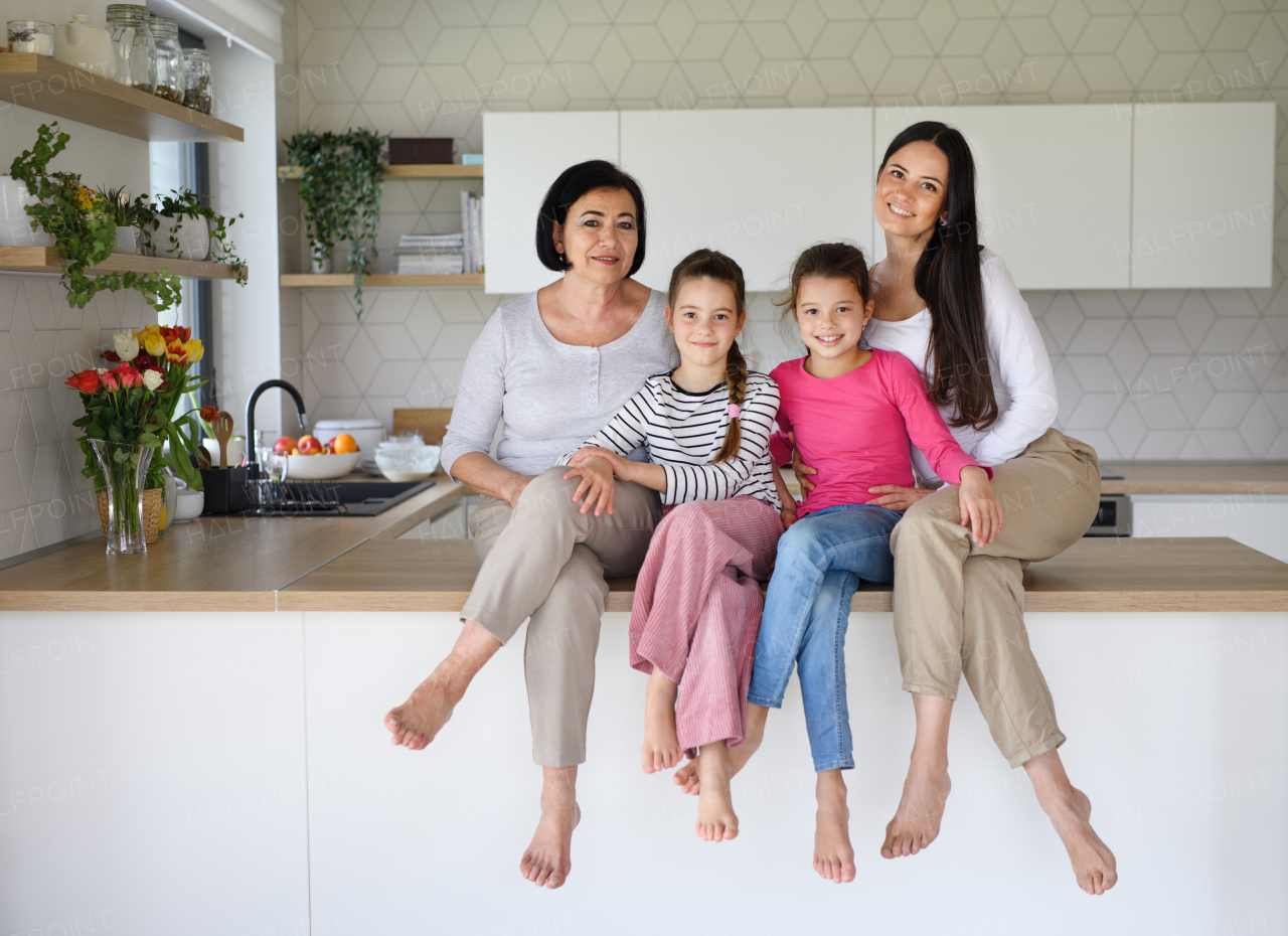 Portrait of happy small girls with mother and grandmother sitting on kitchen counter indoors at home, looking at camera.