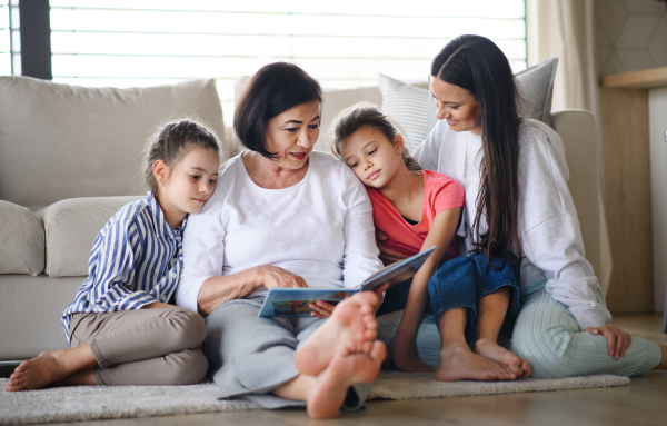 Portrait of happy small girls with mother and grandmother indoors at home, reading story book.