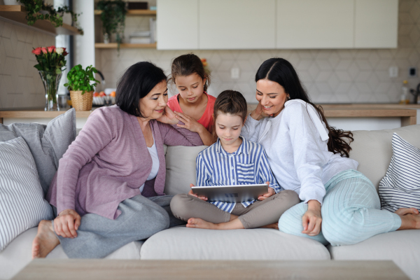 Happy small girls with a mother and grandmother indoors at home, sitting on sofa and using tablet.