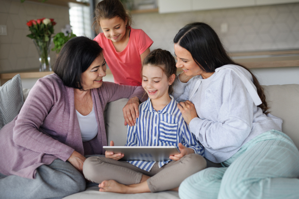 Portrait of happy small girls with mother and grandmother indoors at home, using tablet.