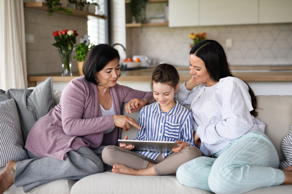 Portrait of happy small girl with mother and grandmother indoors at home, using tablet.