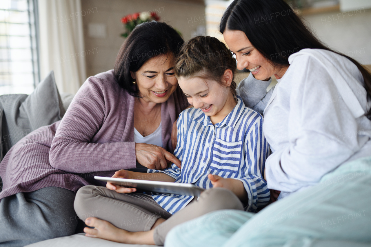 Portrait of happy small girl with mother and grandmother indoors at home, using tablet.