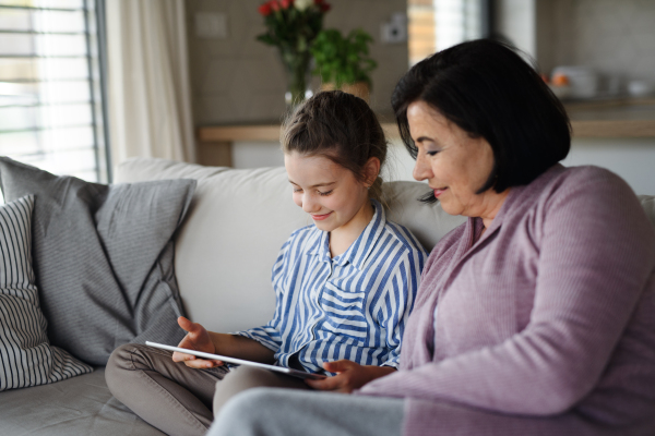 A happy small girl with grandmother indoors at home, using tablet.