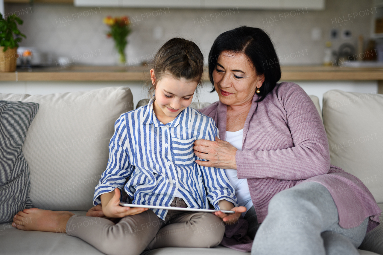 A happy small girl with grandmother indoors at home, using tablet.