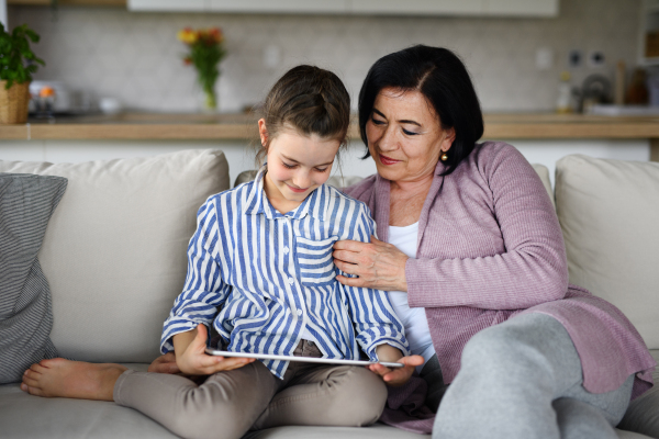 A happy small girl with grandmother indoors at home, using tablet.