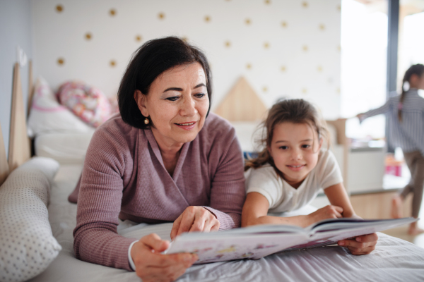 A happy small girl with senior grandmother indoors in bedroom at home, reading on bed.