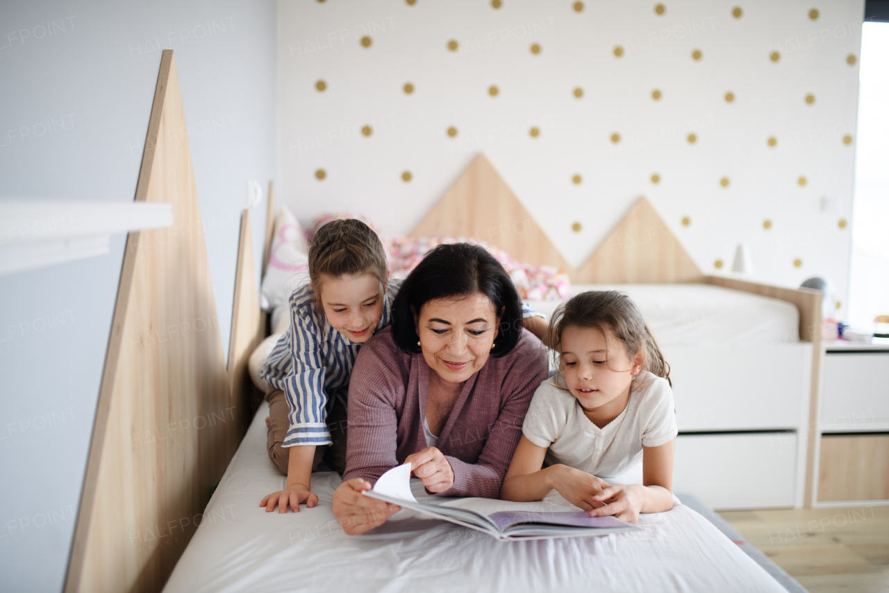 Happy small girls with a senior grandmother indoors in bedroom at home, reading on bed