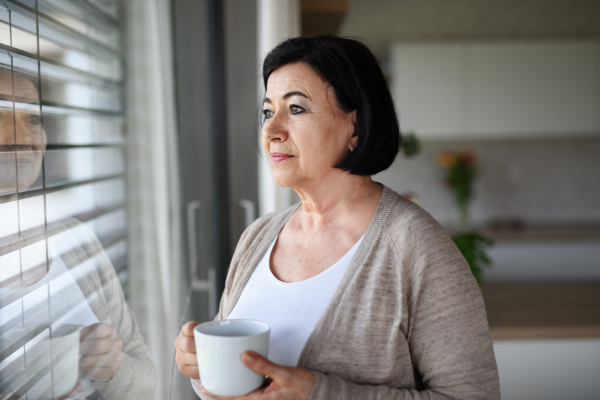 A sad senior woman standing and looking out through window indoors at home.