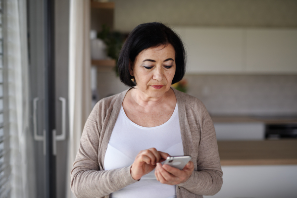 A senior woman standing and using smartphone indoors at home.