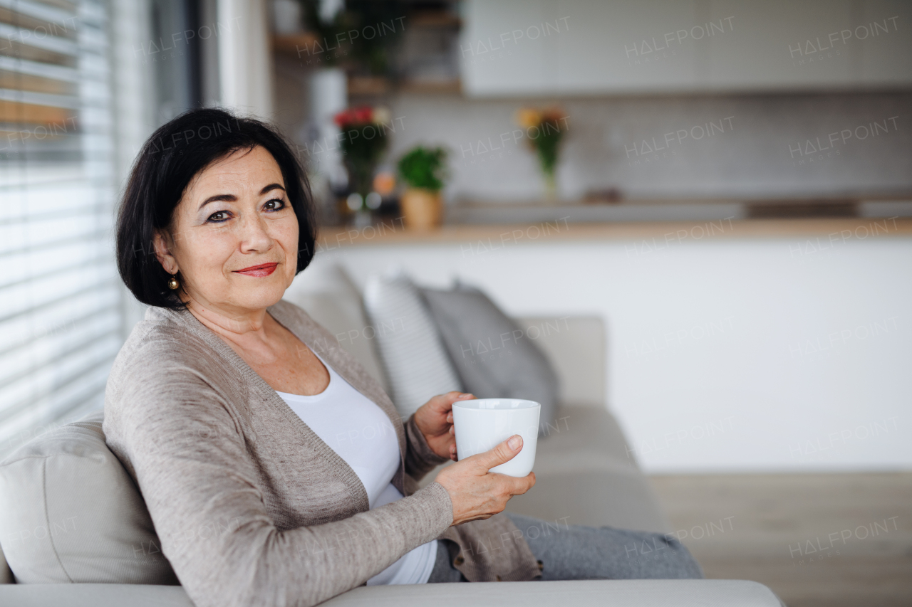 A happy senior woman with coffee sitting on sofam and looking at camera indoors at home.