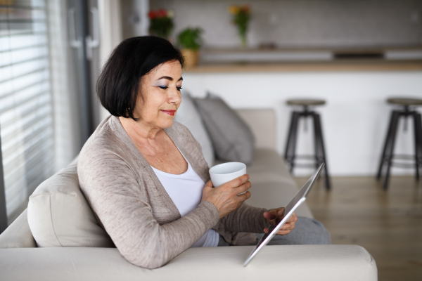 A happy senior woman with tablet sitting on sofa indoors at home, video call concept.