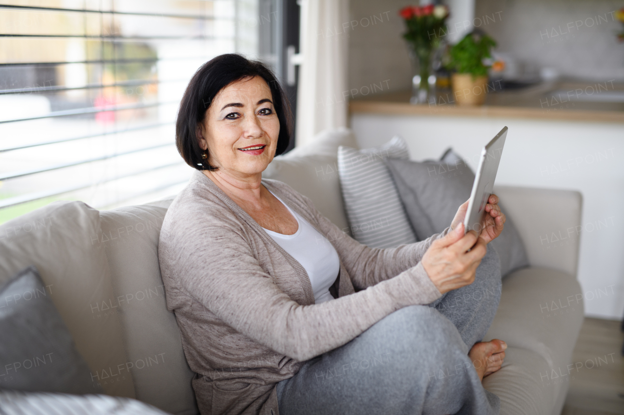 A happy senior woman with tablet sitting on sofa and looking at camera indoors at home, video call concept.