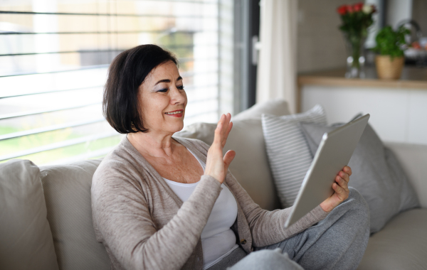 A happy senior woman with tablet sitting on sofa indoors at home, video call concept.