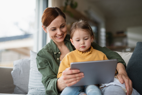 A happy mother with small daughter watching kids programme on tablet indoors at home, single parenting concept.