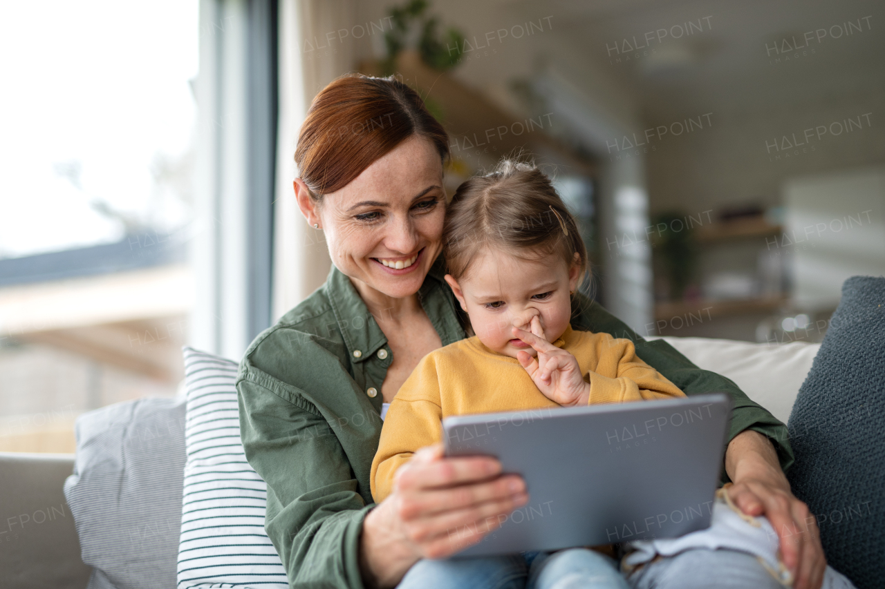 A happy mother with small daughter watching kids programme on tablet indoors at home, single parenting concept.