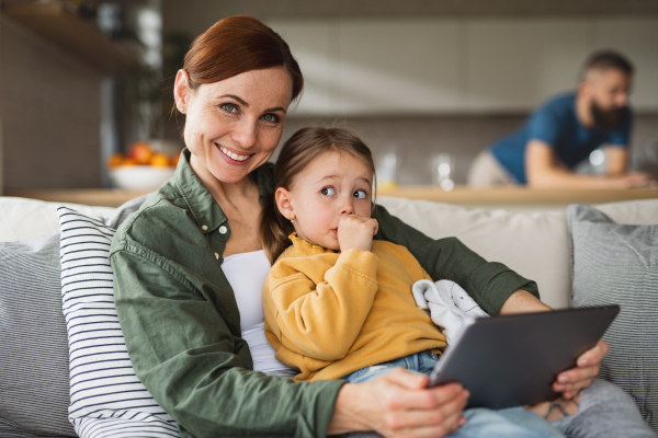 A mother with small daughter using tablet indoors at home, looking at camera, everyday life and home office with child concept.