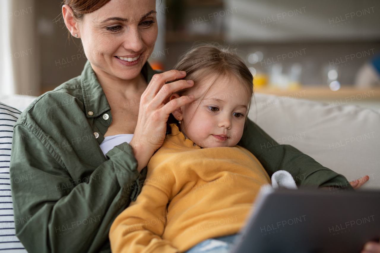 A happy mother with small daughter watching kids programme on tablet indoors at home, single parenting concept.